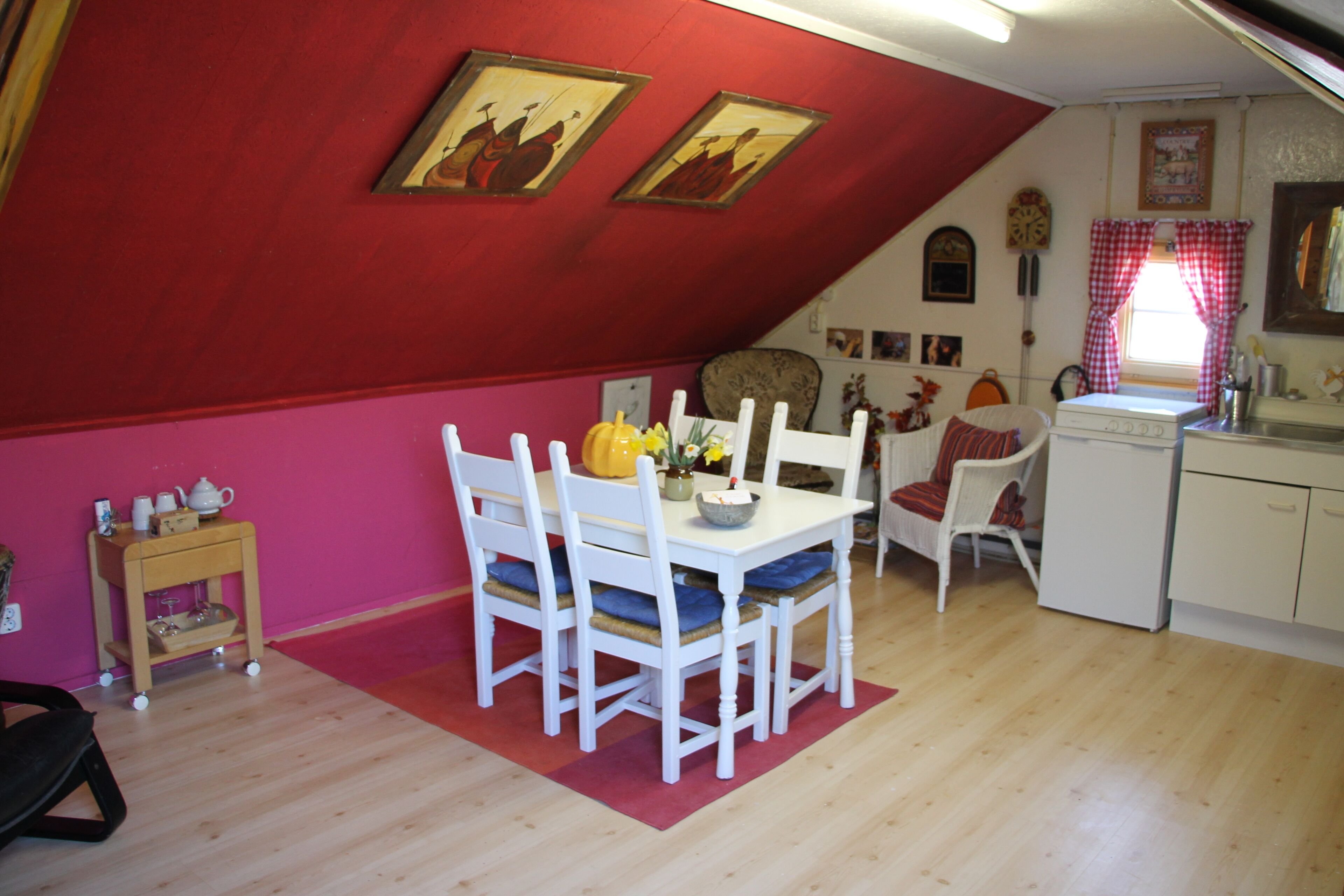 Ein Essbereich mit einem Holztisch und Stühlen, einem kleinen Beistelltisch mit Teekanne sowie einer roten Wand mit zwei gerahmten Bildern.