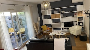 A modern living room with a black sofa, white armchairs, a glass door leading to a balcony, and a white bookshelf.