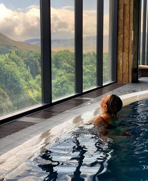 Une personne se détend dans un bain à remous, avec vue sur un paysage boisé grâce à de grandes fenêtres.
