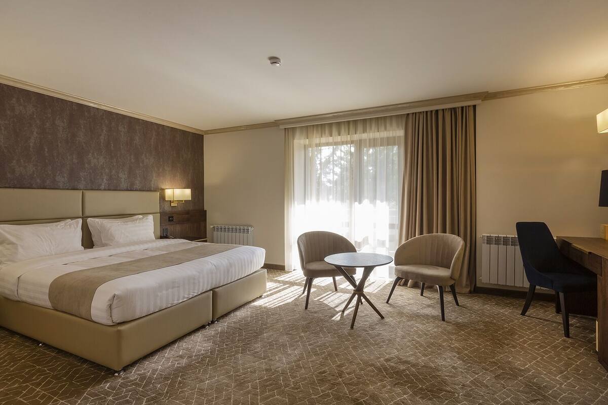 A hotel room with a large bed, two armchairs, a small round table, a desk, and a window with curtains.