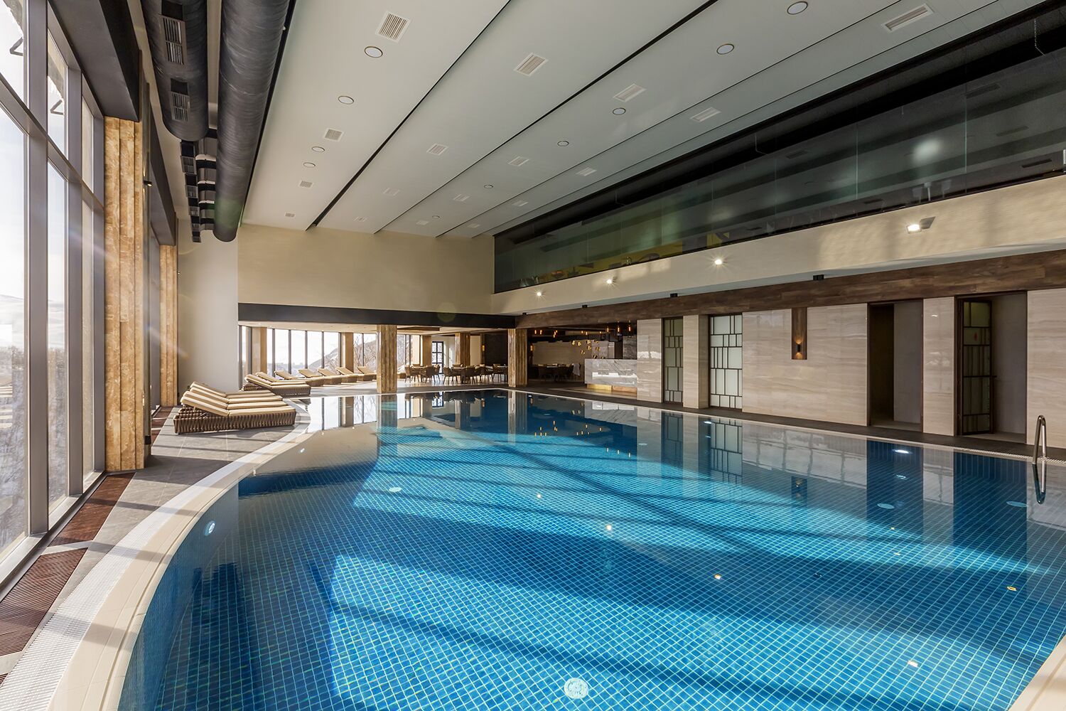 Une grande piscine intérieure avec un sol carrelé, baignée de lumière grâce à de grandes fenêtres, et entourée d’espaces de détente et d’un bar.