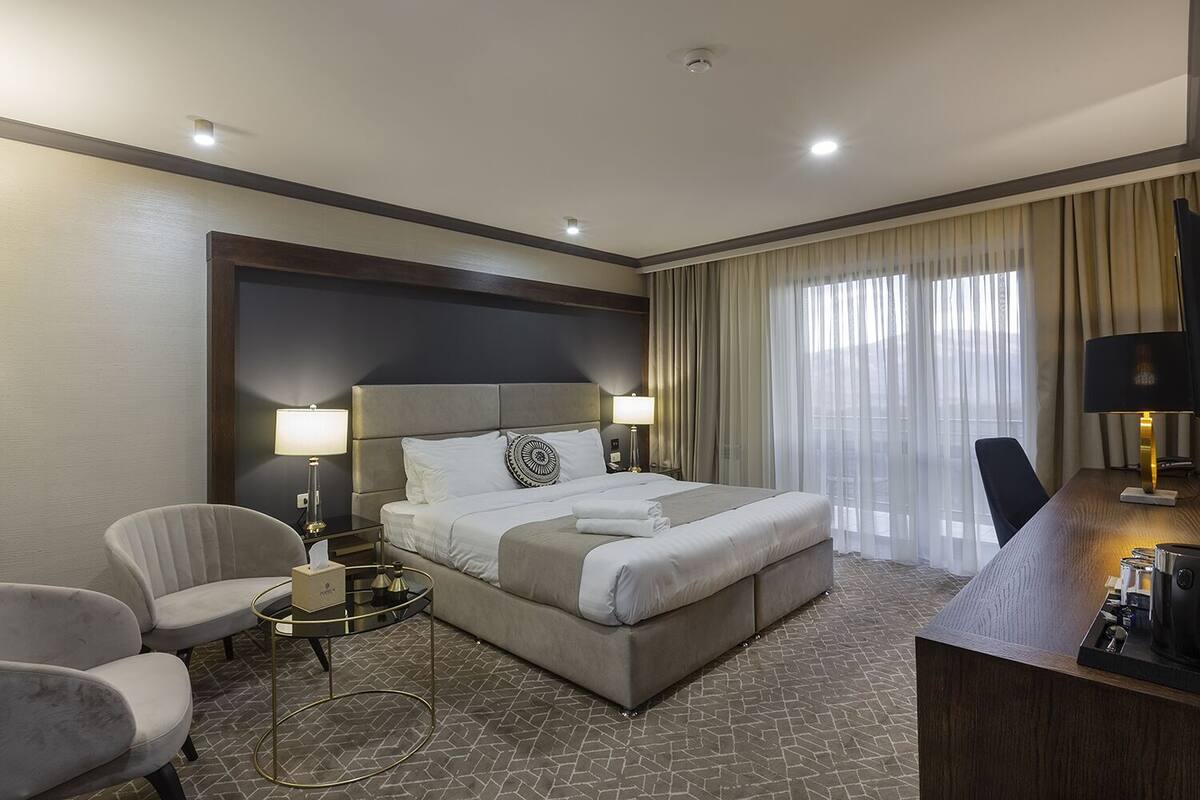 A hotel room with a bed, two bedside tables with lamps, a chair, a desk with a lamp, and a view of the outside through large windows.