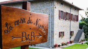 Edessä on kivirakennus, jossa on punaiset ikkunaluukut ja portaikko. Rakennuksen edessä on puinen kyltti, jossa lukee ”La casa di Nino - B&B”, ja nuoli osoittaa oikealle.