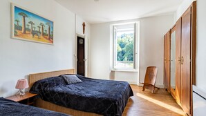 A bedroom with two beds, a wooden wardrobe, a window with a view of trees, and a painting on the wall.