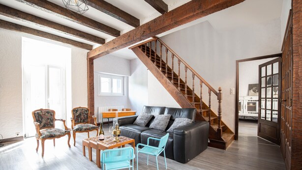 A spacious living room with a black leather sofa, two patterned armchairs, a wooden coffee table, and a blue metal chair. A wooden staircase with a railing leads to another room.