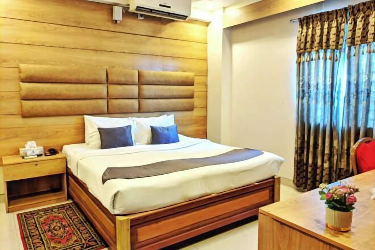 A bedroom with a wooden headboard, a bed with white bedding, a nightstand, a desk with a vase of flowers, and a window with curtains.