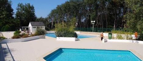 Zona de piscina con un pequeño edificio, rodeada de árboles y una zona vallada.