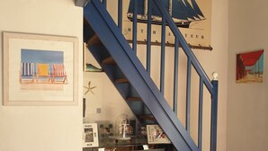 Eine Treppe mit blauen Geländern führt zu einer Empfangsebene mit einem Samsung Fernseher, gerahmten Kunstwerken und einem kleinen Tisch mit einer Tischdecke im Seestil.