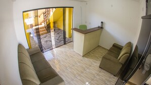 A compact living space with a sofa, a small kitchenette, and a sliding glass door leading to a balcony. - Pousada Sol de Maria (Penha)