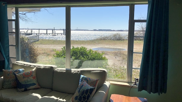 A room with a large window overlooking a beach, a sofa with decorative pillows, and a small wooden table. - Fisherman's Cabin on the Bay (Norfolk)