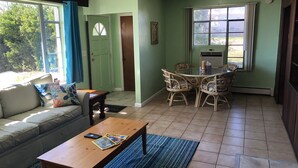 A living room with a sofa, coffee table, and dining area with chairs. There is a door leading outside, a window with curtains, and a radiator. - Fisherman's Cabin on the Bay (Norfolk)
