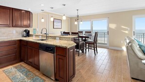 A kitchen with a large island, wooden cabinets, and a dishwasher. There is a dining area with a table and chairs, and a view of the ocean through large windows.