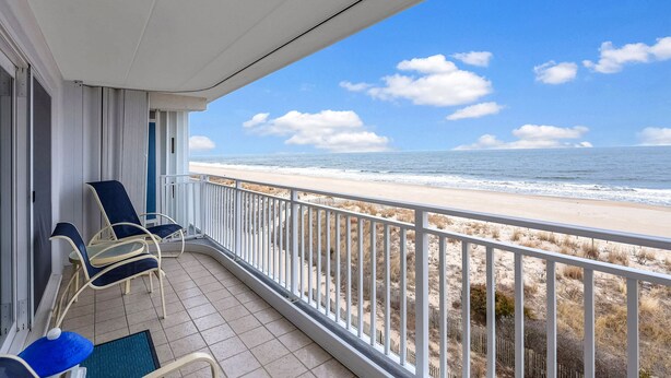 A balcony with two blue chairs and a small table, overlooking a beach with a fence and the ocean.