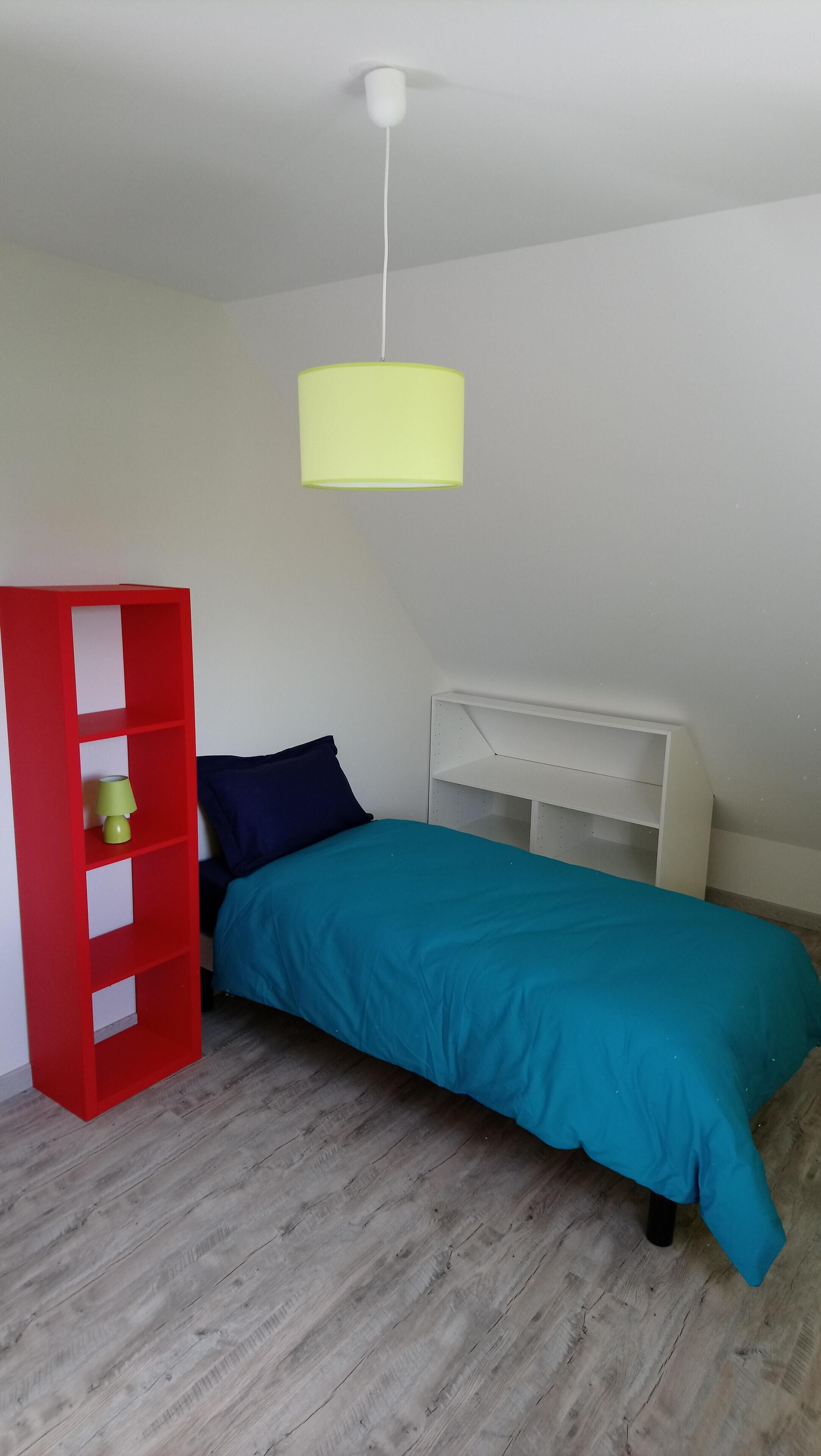 A bedroom with a teal bedspread, a red bookshelf, a white nightstand, and a yellow hanging light fixture.