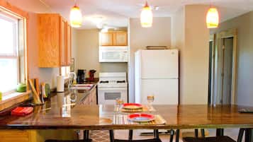 Una cocina con armarios de madera, microondas, refrigerador y un comedor con mesa y sillas de vidrio.