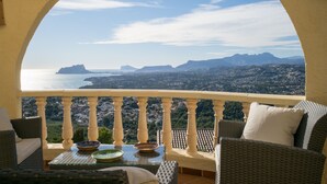 Un balcón con mobiliario de mimbre, una mesa de vidrio y vistas al mar y las montañas.