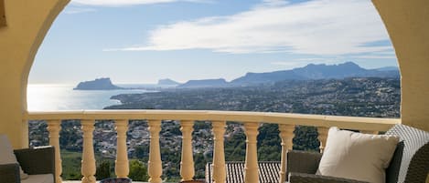 A balcony with wicker furniture, a glass table, and a view of the sea and mountains.