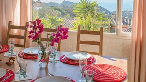 Mesa para dos servida con platos rojos y blancos, vasos transparentes y un florero con flores rosas. La mesa está situada cerca de una ventana con vistas a un paisaje costero con colinas y el mar.