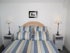 Uma cama bem arrumada com roupa de cama listrada, duas mesas de cabeceira com abajures, um quadro na parede e uma cabeceira de vime.