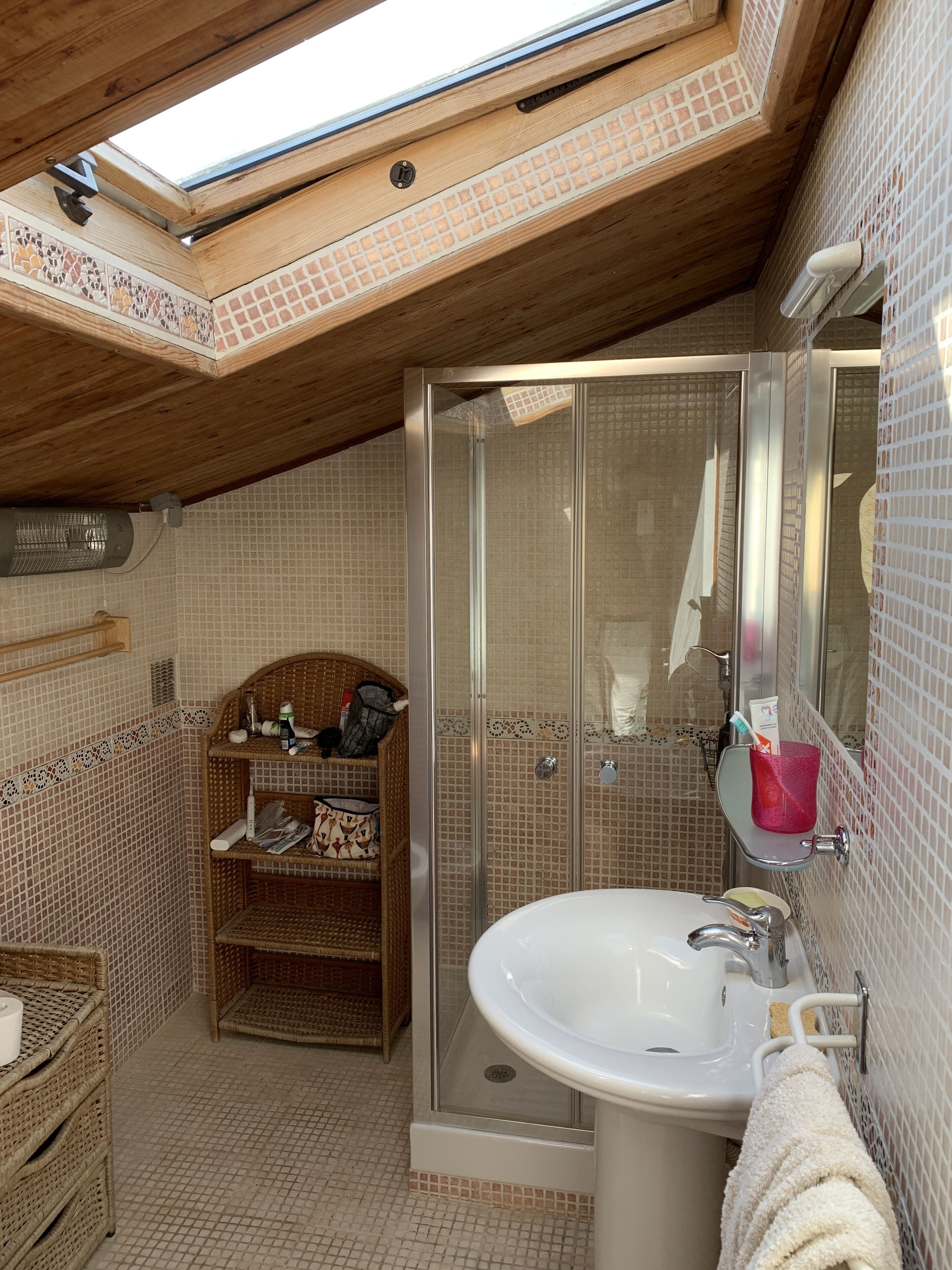 A bathroom with a skylight, a glass shower enclosure, a white sink, and a wicker shelf with toiletries.