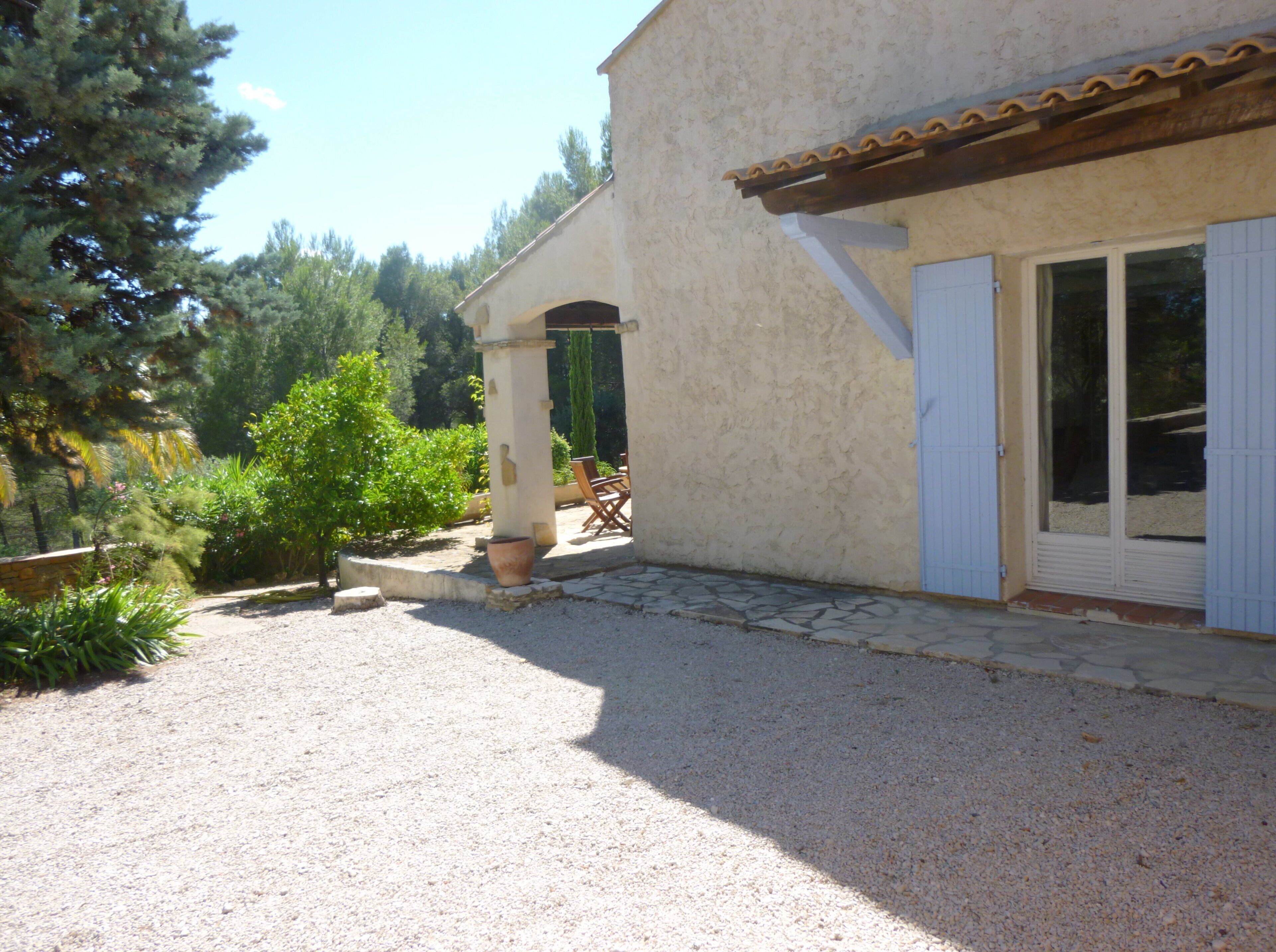 Un bâtiment avec une allée gravillonnée, un patio couvert avec des chaises, et une vue sur les arbres et la verdure.
