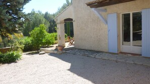 Un bâtiment avec une allée gravillonnée, un patio couvert avec des chaises, et une vue sur les arbres et la verdure.