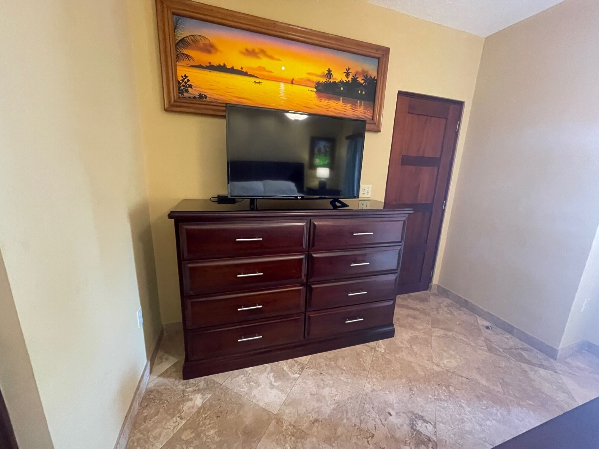 A room with a dark wooden dresser, a flat-screen TV on top, and a framed painting of a sunset over water.
