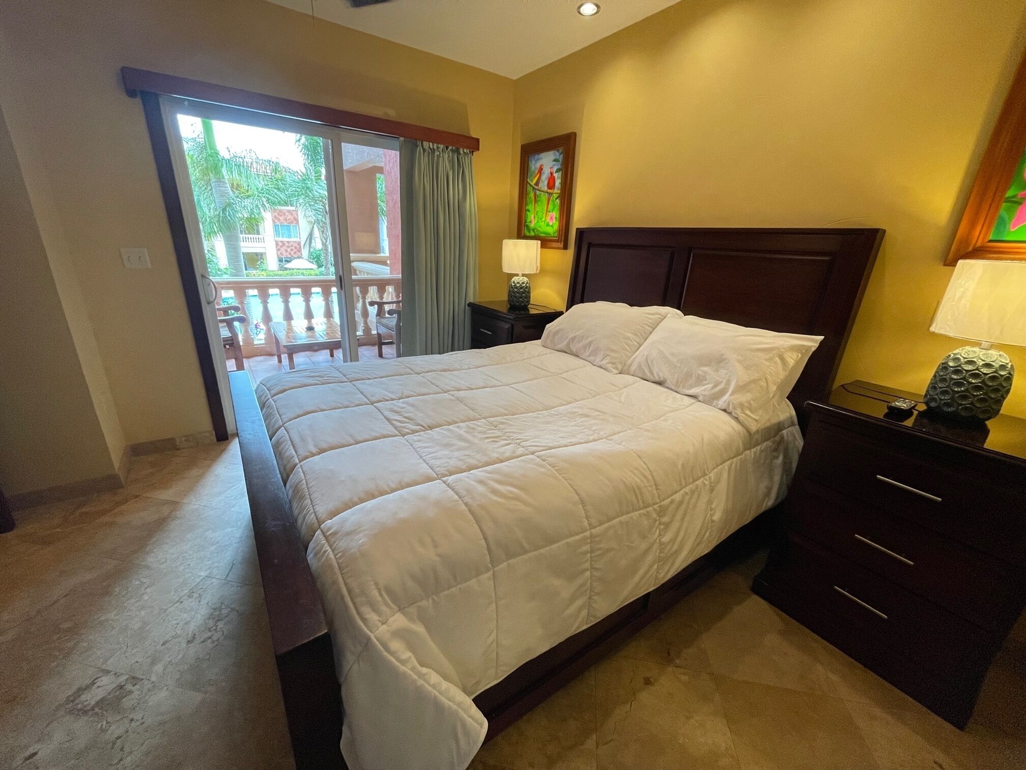 Habitación de hotel con una cama grande, una mesita de noche con lámpara, un cuadro en la pared y vistas al balcón exterior.