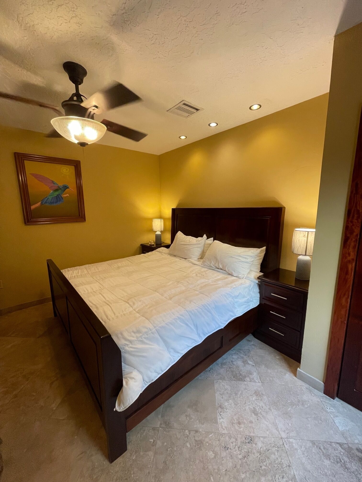A bedroom with a bed, nightstands, a ceiling fan, recessed lighting, and a wall-mounted painting.