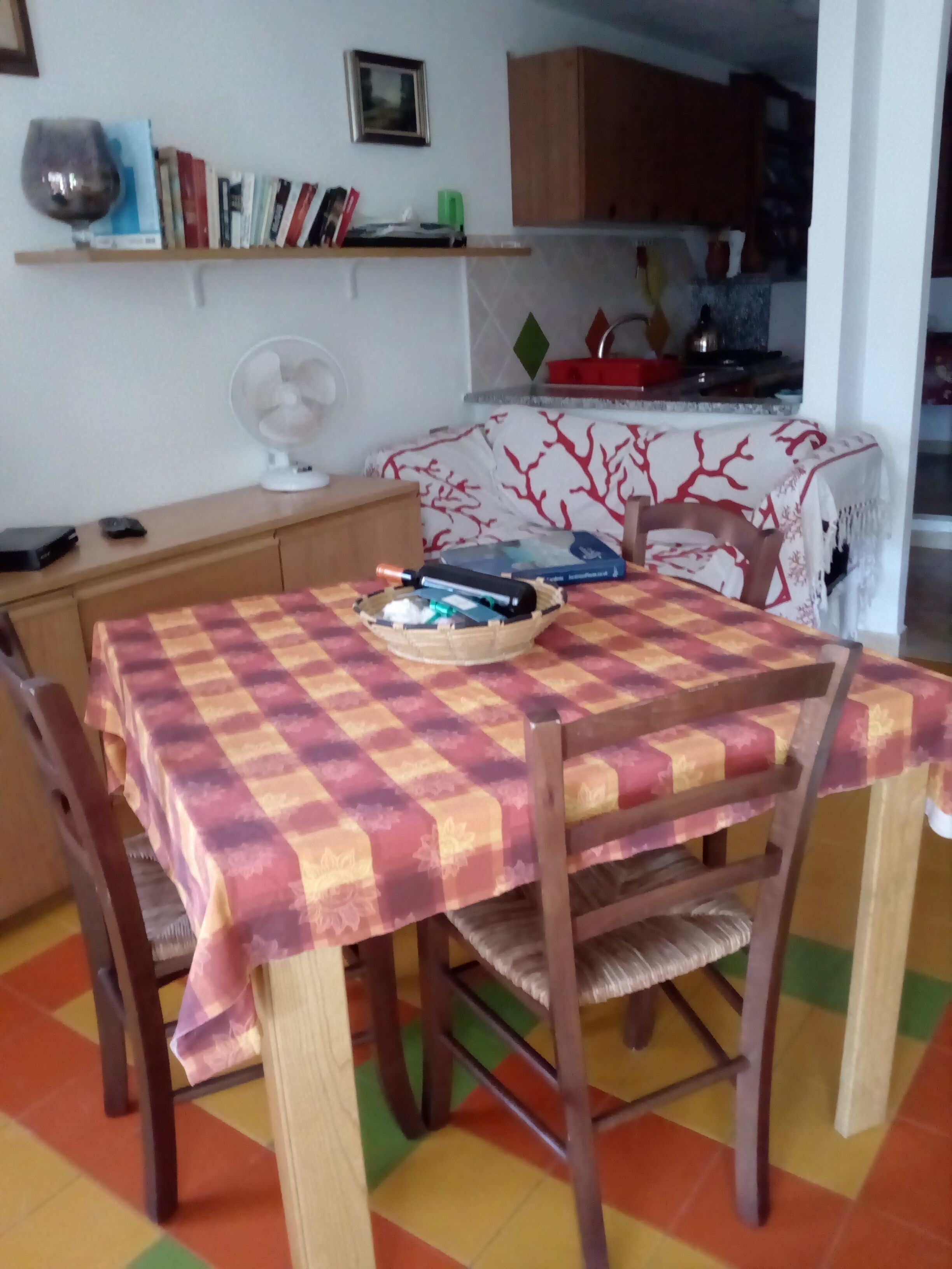 A dining area with a checkered tablecloth, wooden chairs, and a fan. A bookshelf with books and a wine glass is visible in the background.