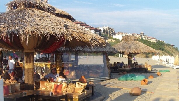 A beachside restaurant with thatched umbrellas, outdoor seating, and a view of the beach and buildings in the background.