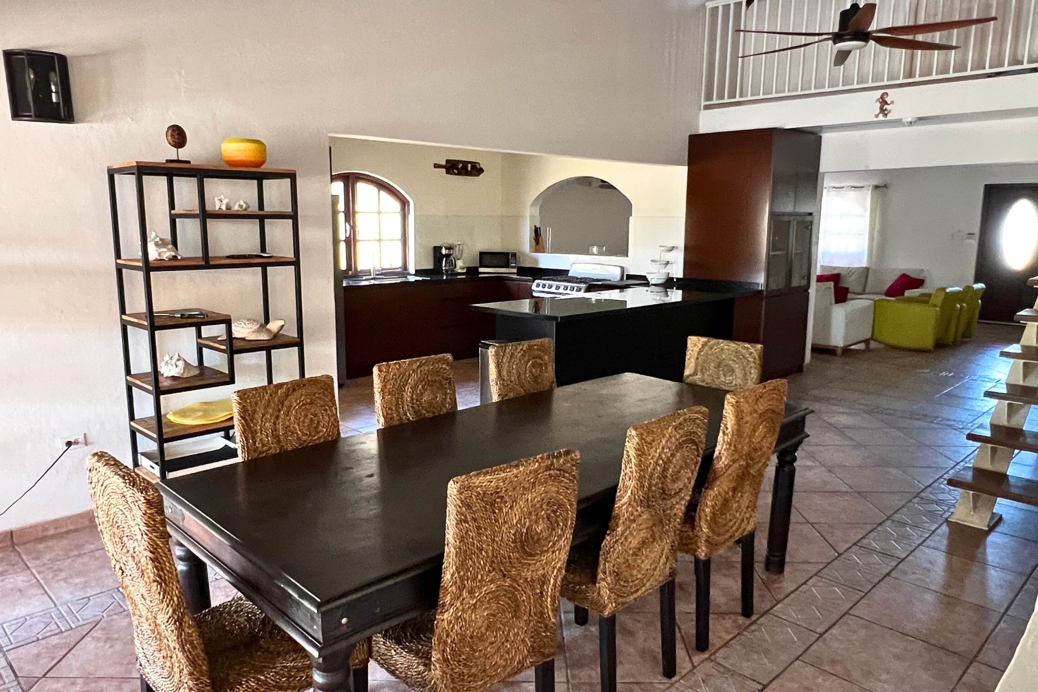 A dining area with a dark wooden table and woven chairs, a kitchen in the background, and a staircase leading to an upper level.