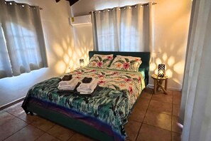 Una cama con una colcha estampada, dos toallas, una mesita de noche con una lámpara y cortinas.