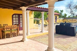 Un espacio de patio cubierto con una mesa, sillas, una parrilla y un edificio amarillo al fondo.