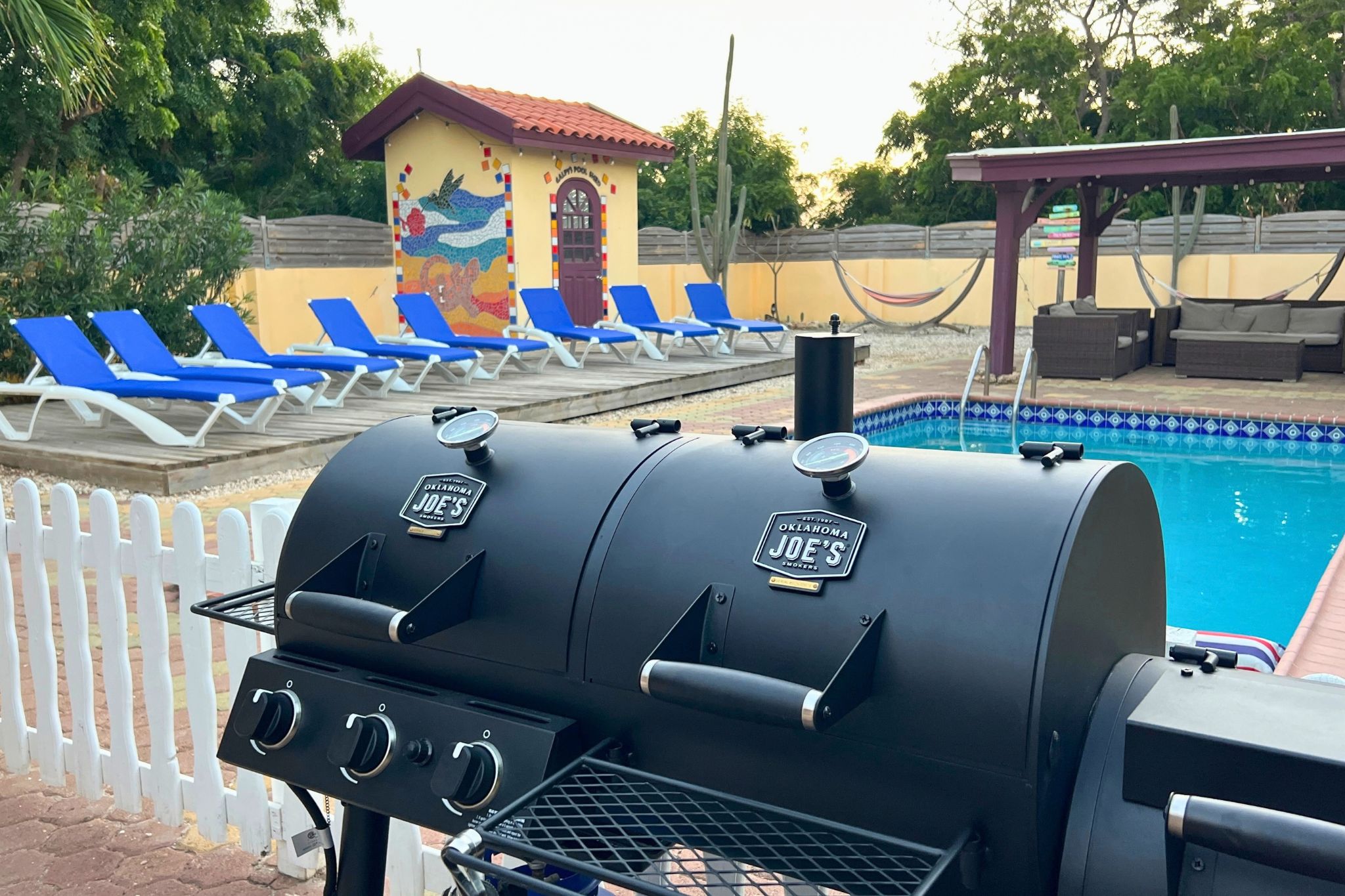 Un barbecue arborant la marque « OKLAHOMA JOE’S », une piscine aux carreaux bleus et un petit bâtiment orné d’une fresque colorée.