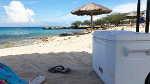 Eine Strandansicht mit einem Reetdachschirm, einer Coleman-Kühlbox und einem Paar Flip-Flops im Sand.