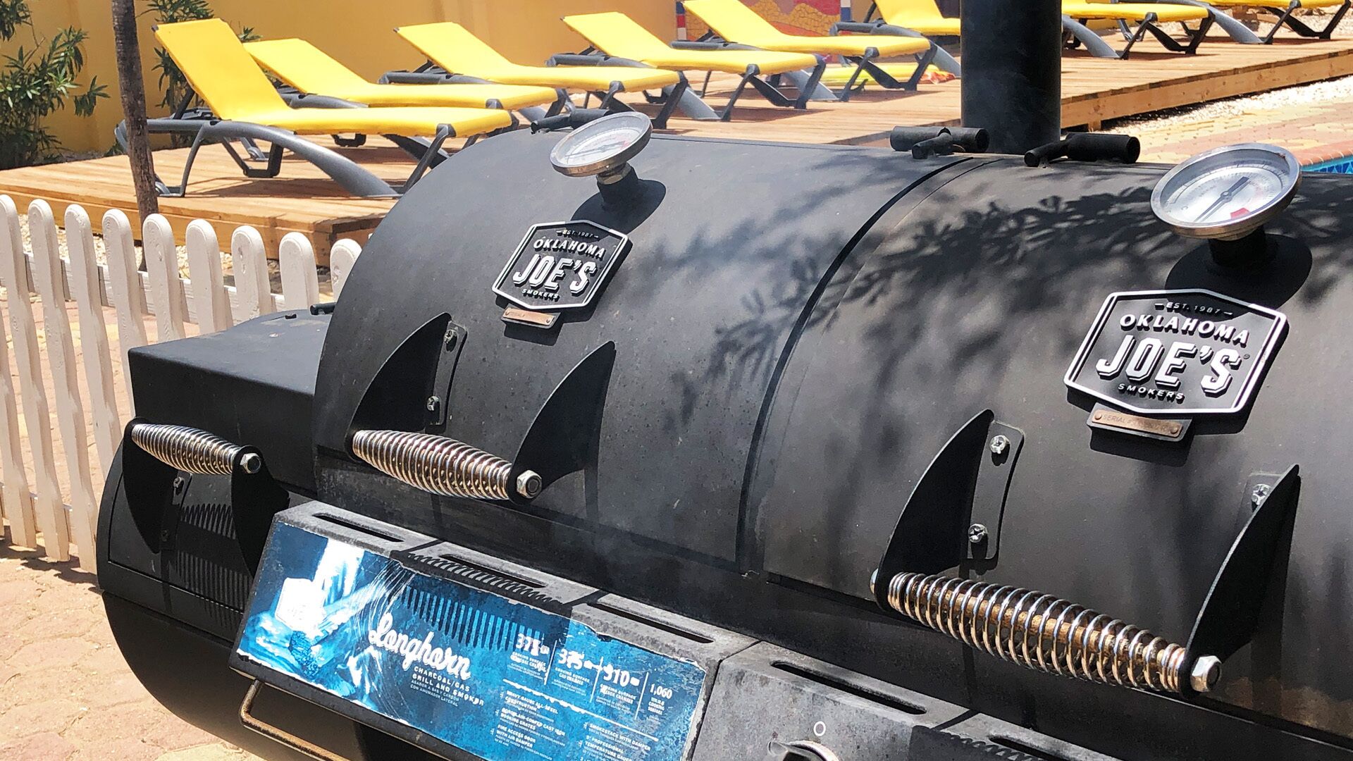 Ein Grill mit "Oklahoma Joe's" Branding, draußen gelegen, im Hintergrund ein Pool und Liegestühle.