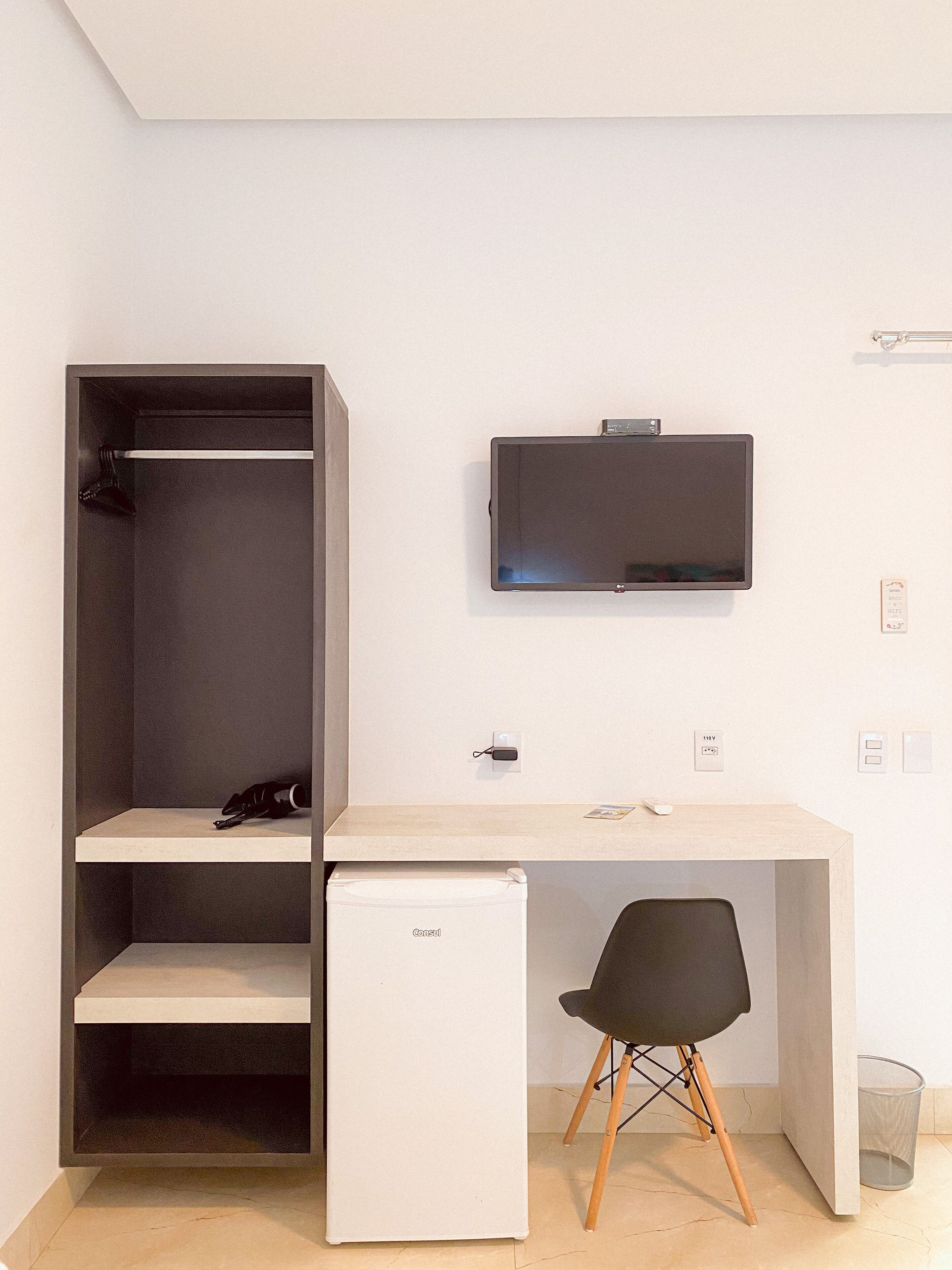 A compact room with a white desk, a small refrigerator, a black chair, and a mounted TV.