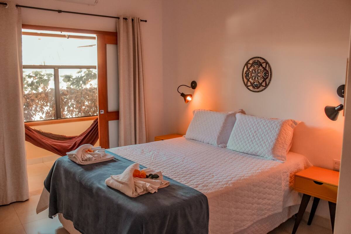 A hotel room with a bed, a nightstand, a wall-mounted lamp, a window with curtains, and a hammock.