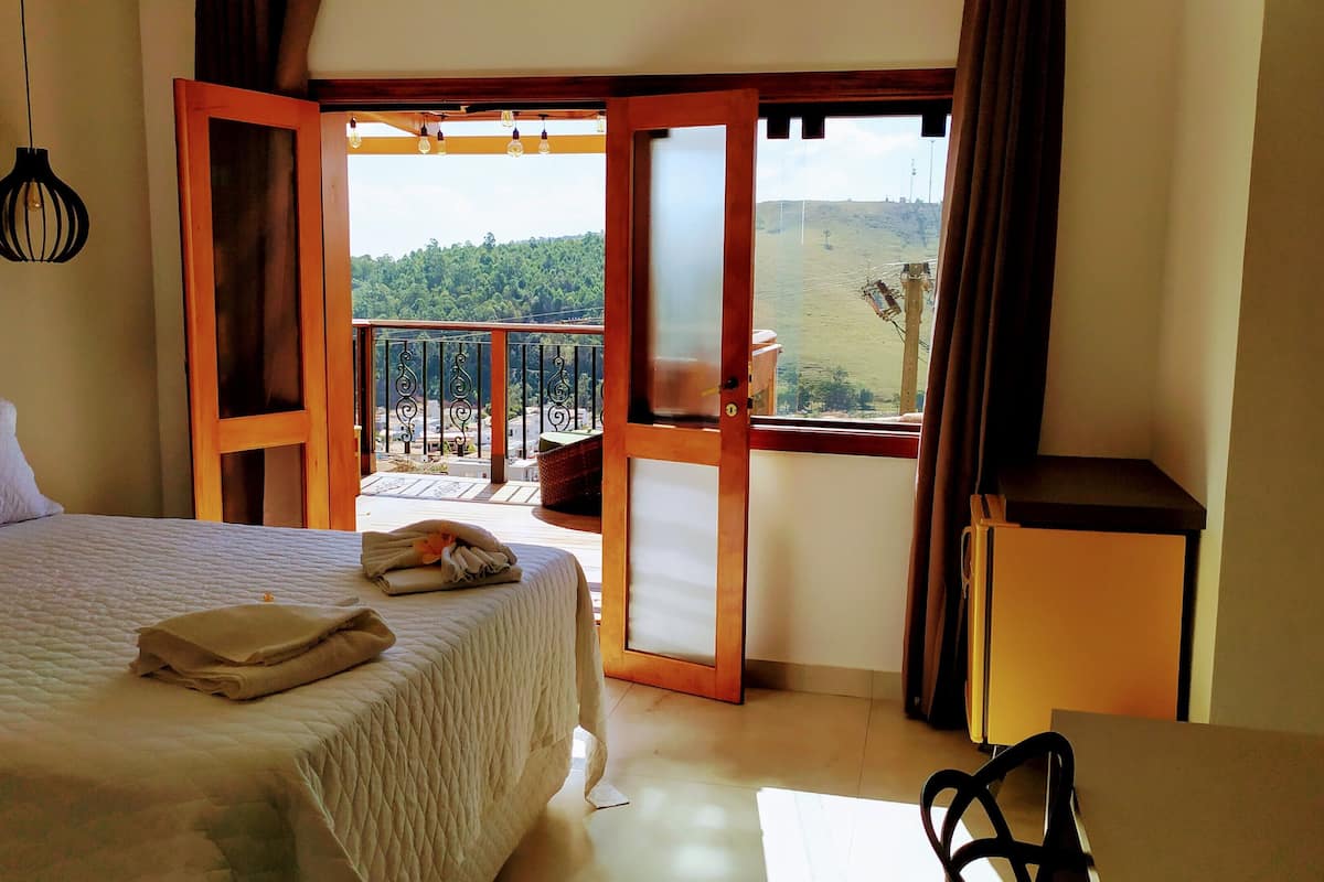 A bedroom with a bed, a bedside table, a chair, and an open door leading to a balcony with a view of the countryside.