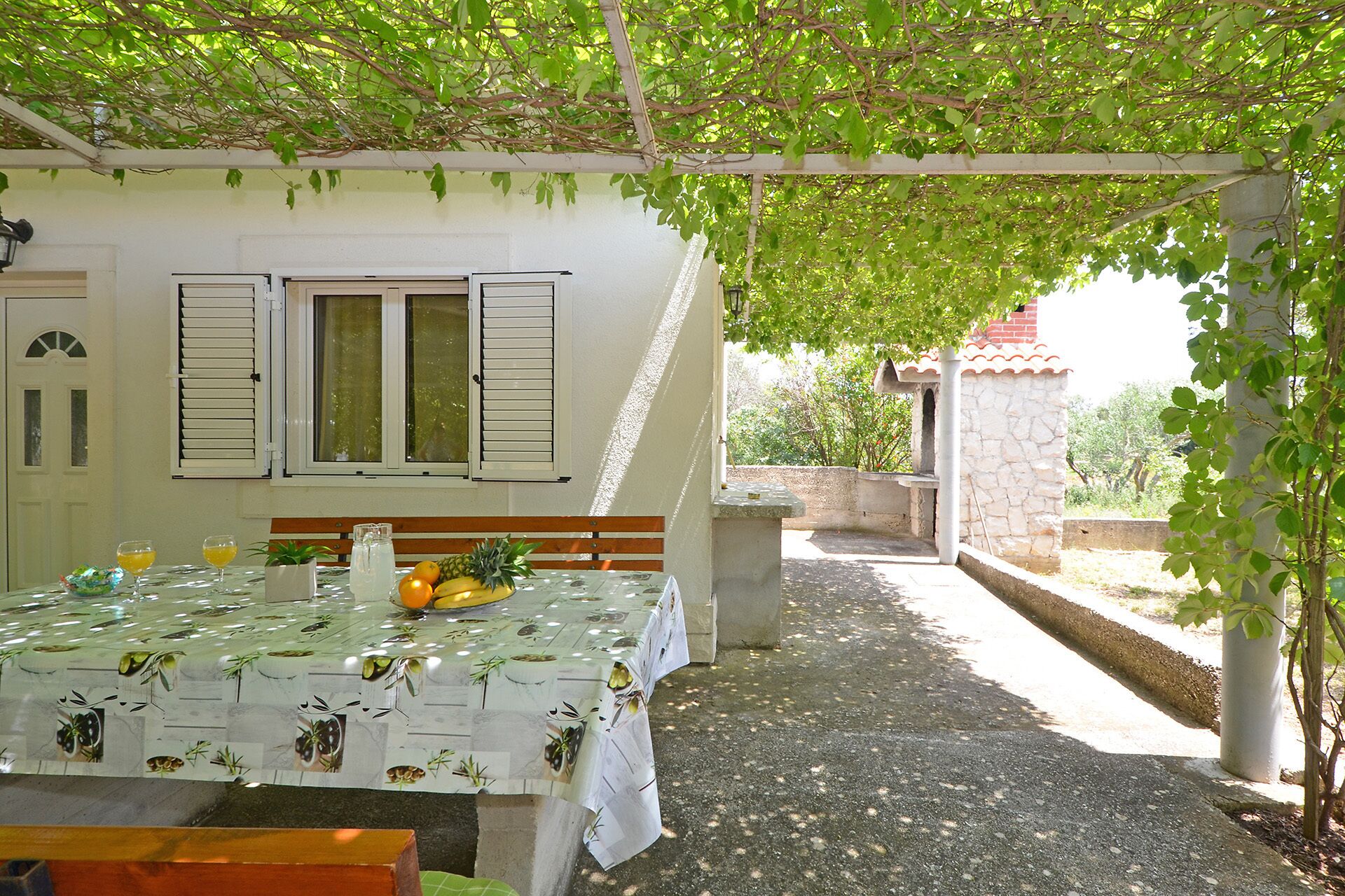 A covered outdoor dining area with a table set for two, a bench, and a view of a house with shutters.