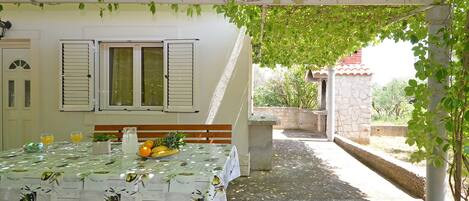A covered outdoor dining area with a table set for two, a bench, and a view of a house with shutters.