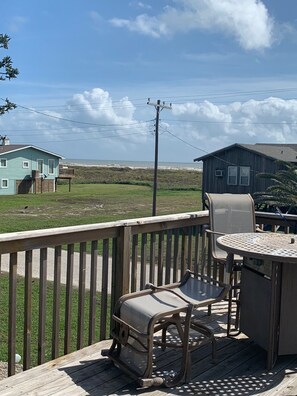 A deck with outdoor furniture, a view of the ocean, and a wooden house in the background.