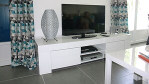 A modern living room with a flat-screen TV on a white entertainment unit, a decorative silver vase, and patterned curtains.