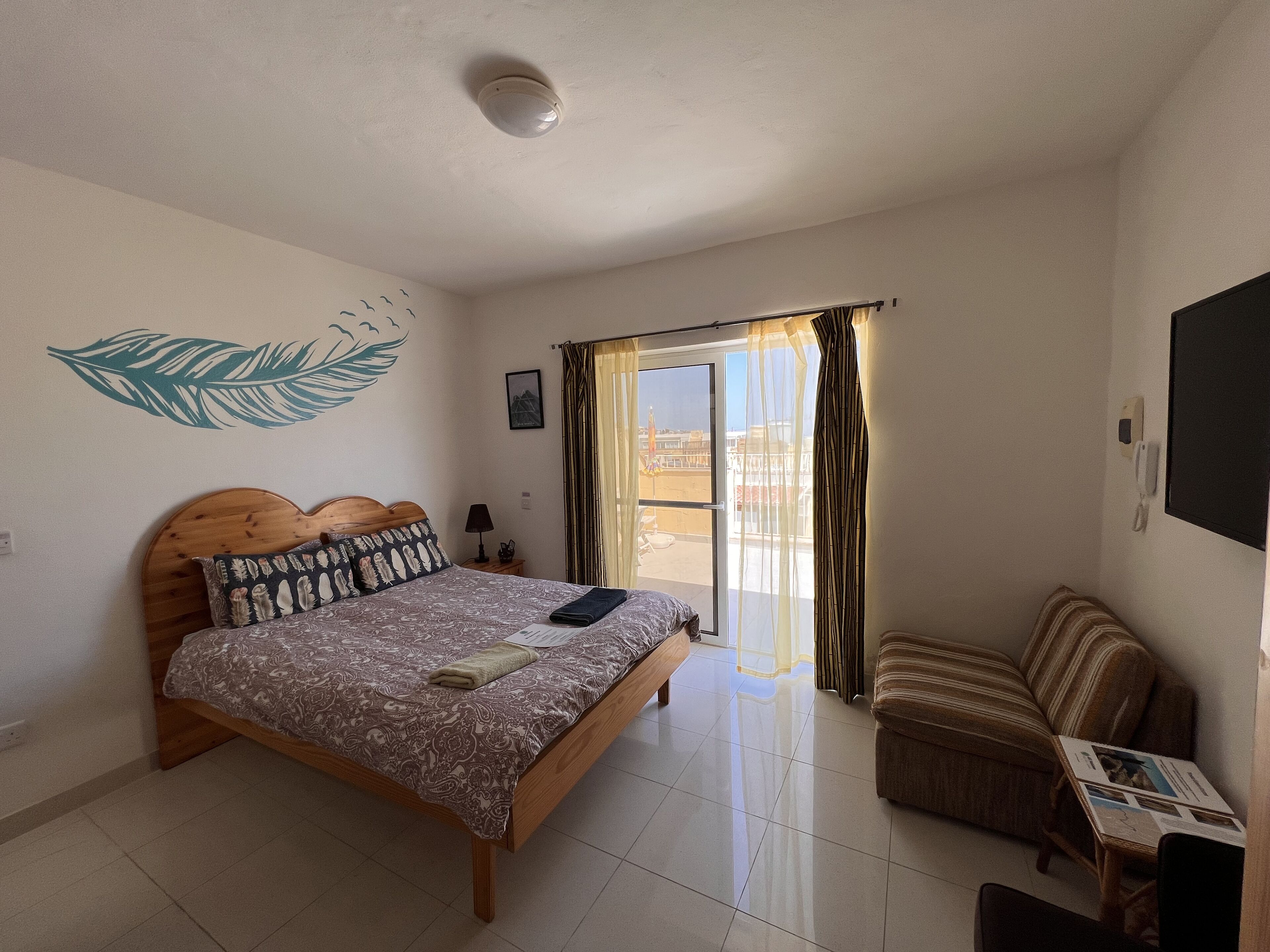 Une chambre à coucher comprenant un lit, une chaise, une table, une fenêtre avec des rideaux, un élément de décoration murale et un luminaire au plafond.