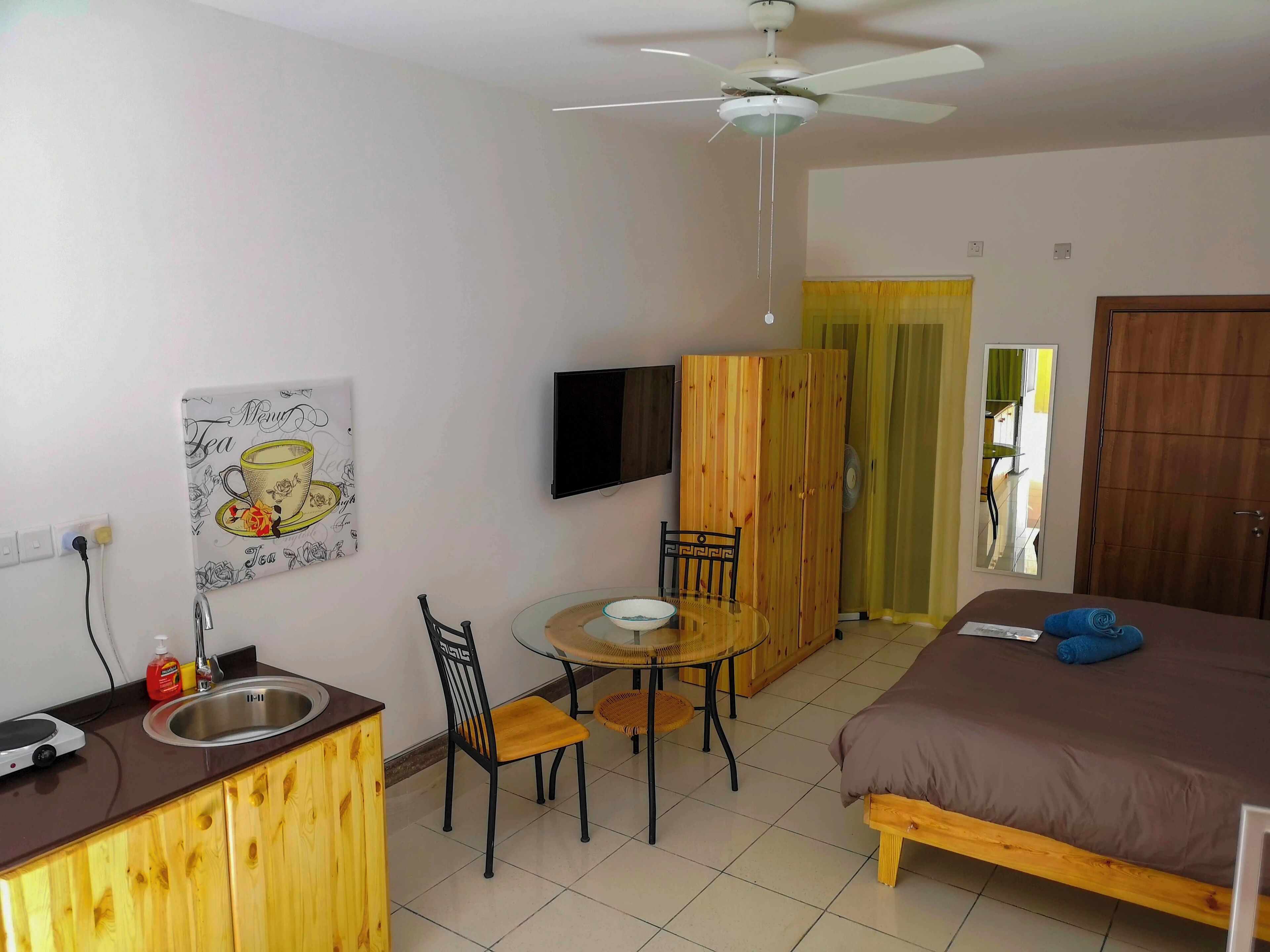 Una habitación de hotel con una cama, una mesita con sillas, un televisor en la pared, un armario de madera y un ventilador de techo.
