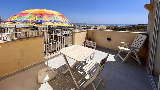 A balcony with a table and chairs, a colorful umbrella, and a view of the sea. - All Nations Holiday Home (Ghajnsielem)