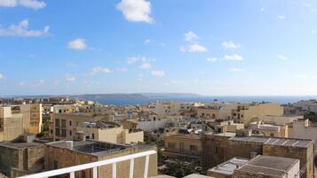 A cityscape with multiple buildings, some with flat roofs and others with sloped roofs, under a clear blue sky with scattered clouds.
