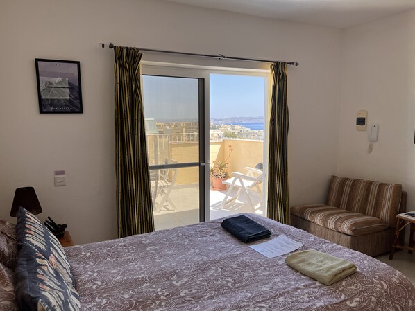 A bedroom with a bed, a chair, a table, a window with curtains, a wall-mounted phone, and a framed picture. - All Nations Holiday Home (Ghajnsielem)