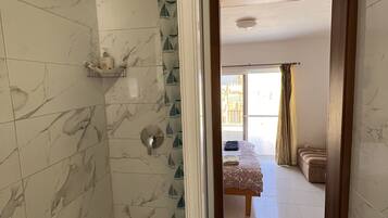 Baño con baldosas de mármol, ducha y váter. Se ve una cama a través de un vano abierto en la habitación contigua.